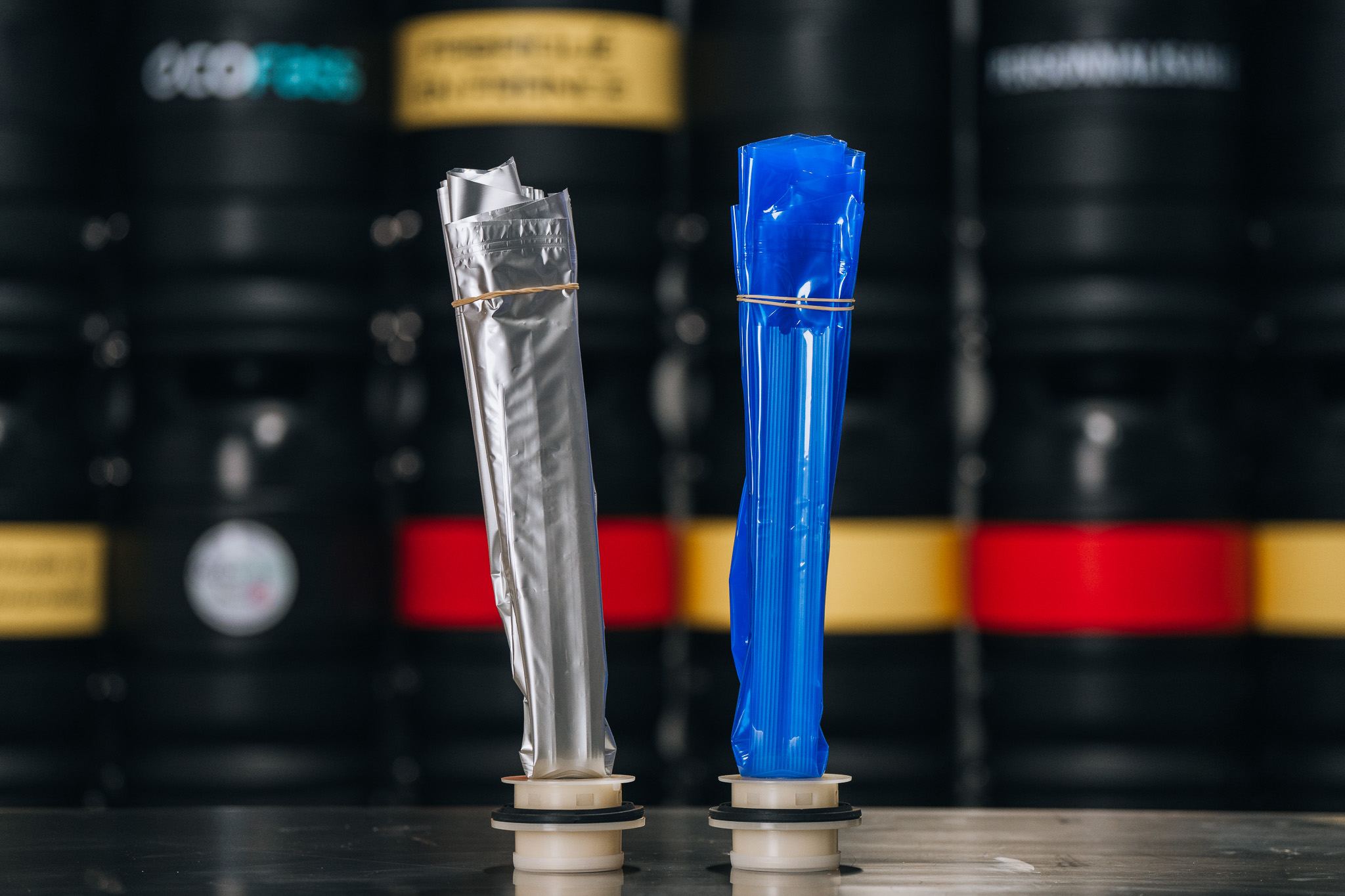 Deux poches ECOFASS dressées sur leur embout, l'une en aluminium, l'autre en plastique bleu, devant des fûts noirs